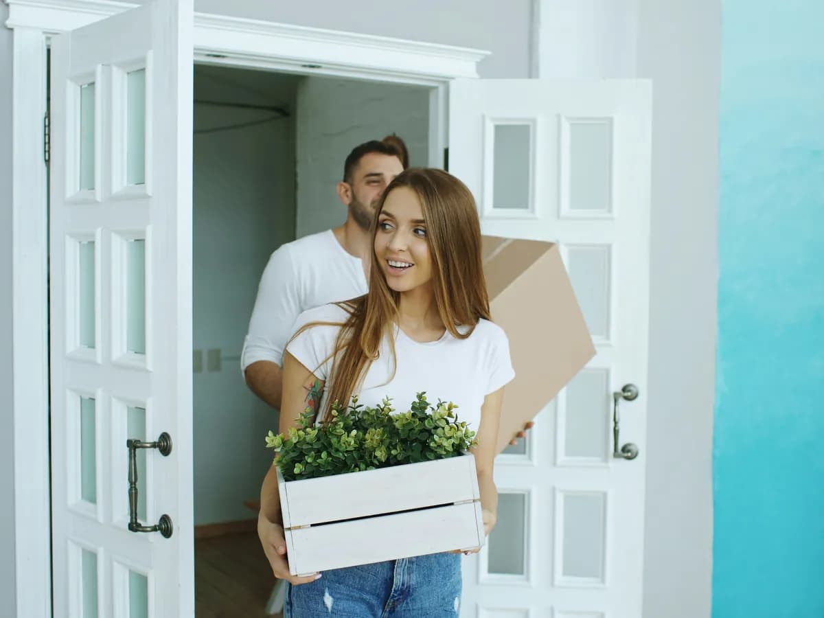 A couple carrying boxes and a plant through the door of their new home